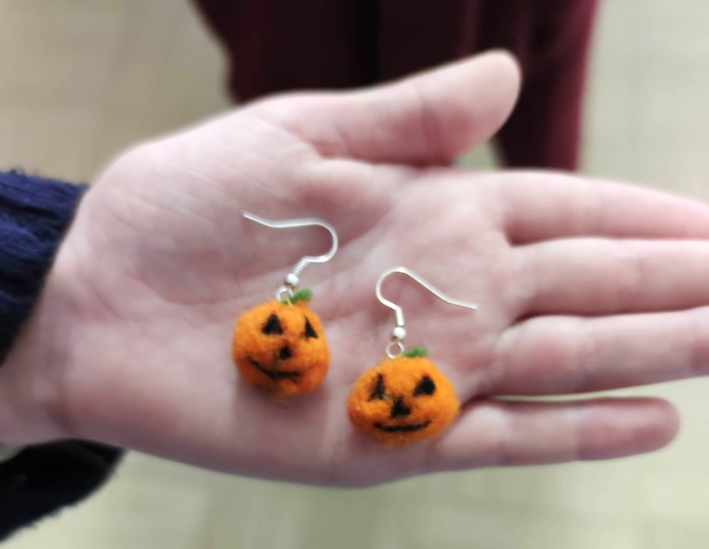 Hand mit zwei orangefarbenen Kürbis-Ohrringen aus Filz, die lachende Gesichter und grüne Blätter haben.