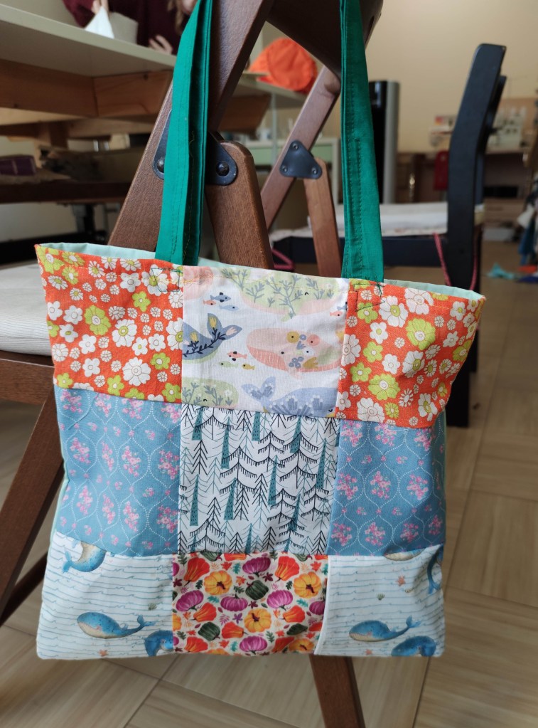 Eine bunte, quadratische Tasche mit verschiedenen Stoffmustern, darunter florale und tierische Designs, hängt an einem Holzstuhl.