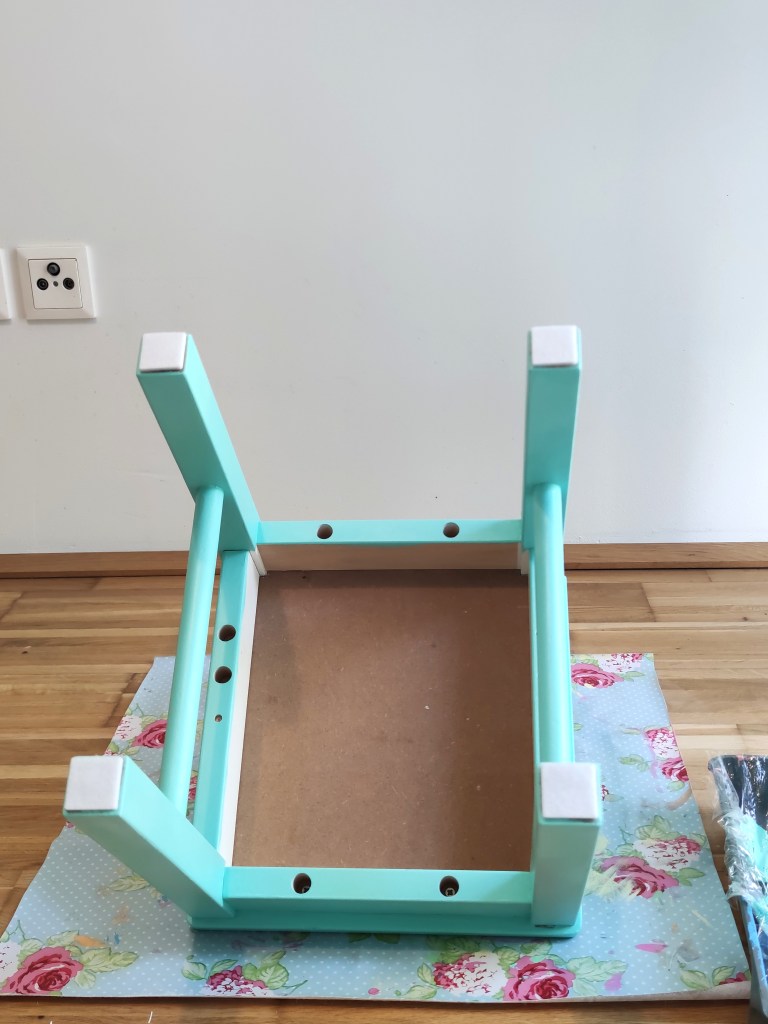Ein türkisfarbener Tischrahmen mit weißen Eckschutz, auf einem blumenmusterigen Tuch auf einem Holzboden liegend.