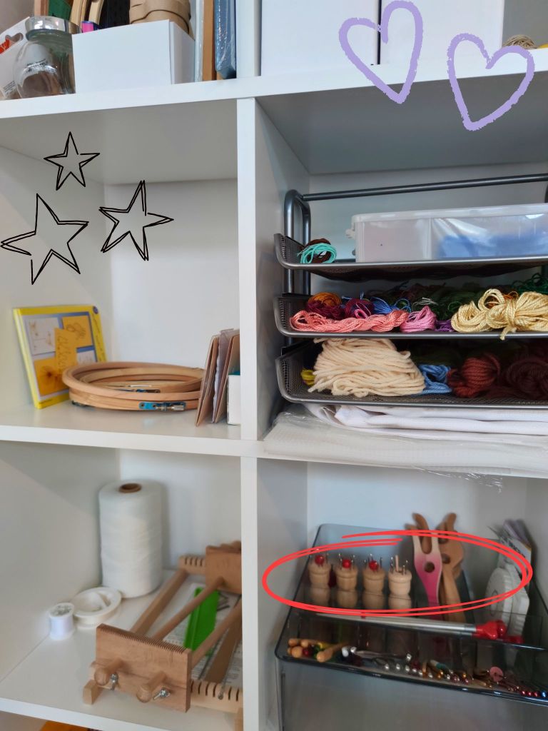 Ein Regal mit verschiedenen Bastelmaterialien, darunter Garn in bunten Farben, Holzrahmen und Werkzeuge. Im Vordergrund sind Holzspulen und weitere Bastelutensilien zu sehen.