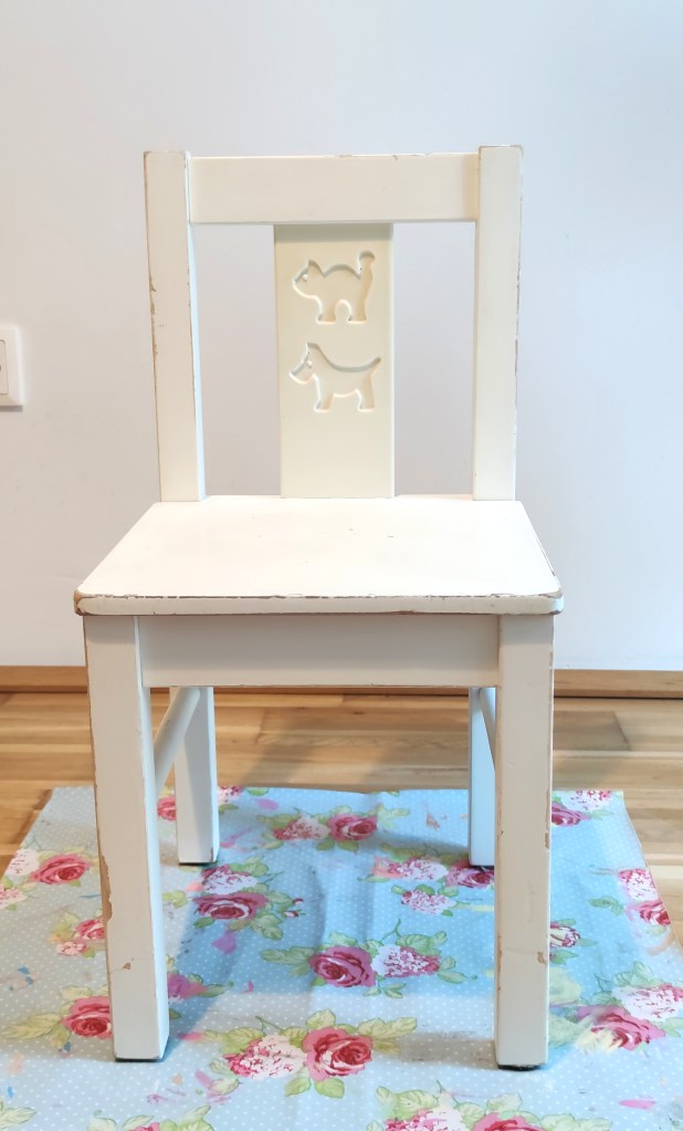 Ein weißer Holzstuhl mit einer Rückenlehne, die zwei ausgeschnittene Tiermotive zeigt, auf einem blauen Teppich mit Blumenmuster.