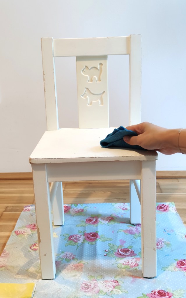 Ein weißer Stuhl mit Tiermotiven auf der Rückenlehne, während eine Hand mit einem blauen Tuch den Stuhl abwischt. Der Stuhl steht auf einem Boden mit einem blumigen Teppich.