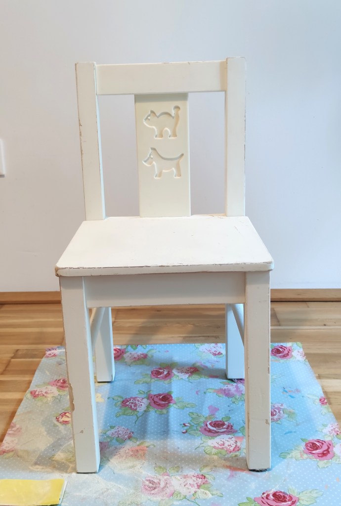 Ein einfacher weißer Stuhl mit einer Rücklehne, die zwei stilisierte Katzensilhouetten zeigt, steht auf einem bunten Untergrund mit Rosenmuster.